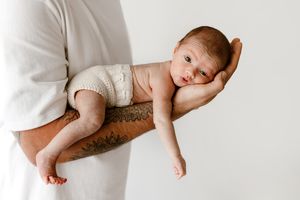 Tattooed father cradling sleeping newborn baby on one arm against white background.