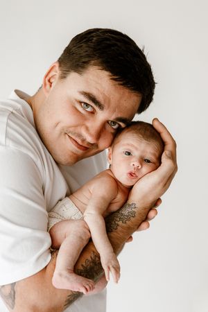 A smiling father with tattoos gently holds his newborn baby against his chest on a white background.