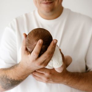 A tattooed father in a white t-shirt gently cradling a newborn baby with dark hair in both hands.