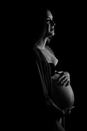Artistic black and white maternity silhouette shows an expectant mother's profile in dramatic lighting against dark background.