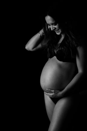 Black and white artistic maternity photo showing a pregnant figure in lingerie against a dark background.