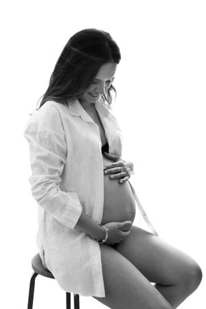 Black and white artistic maternity photo of an expectant mother in a white button-down shirt sitting on a stool.