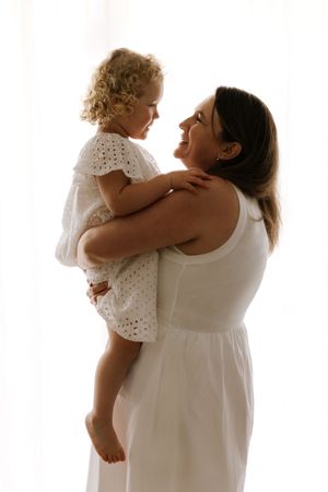 Mother and child wearing white clothing embrace in tender poses against light background.