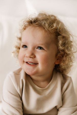 A smiling toddler with curly blonde hair wearing a beige sweater looks up while posing for a portrait.