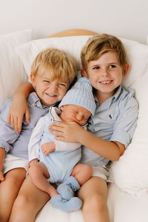 Three siblings in light blue clothing snuggle together on white bedding showing tender affection and closeness.