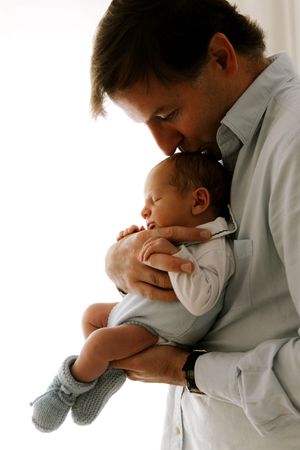 A tender moment captured between a father in white shirt cradling his sleeping infant wearing white onesie and gray booties.