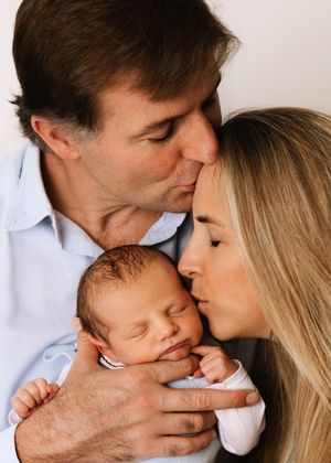A tender family moment as parents lovingly kiss their sleeping newborn baby dressed in white.