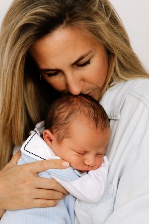 Tender moment captured as mother holds and kisses sleeping newborn baby wrapped in white clothing.