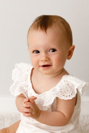 An adorable baby wearing a white eyelet lace dress smiles warmly at the camera against a light background.