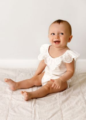 Smiling baby wearing white lace-trimmed outfit sits on light colored textured fabric in studio setting.