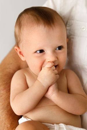 An adorable baby in a white diaper sucking on fingers while resting on a brown surface.