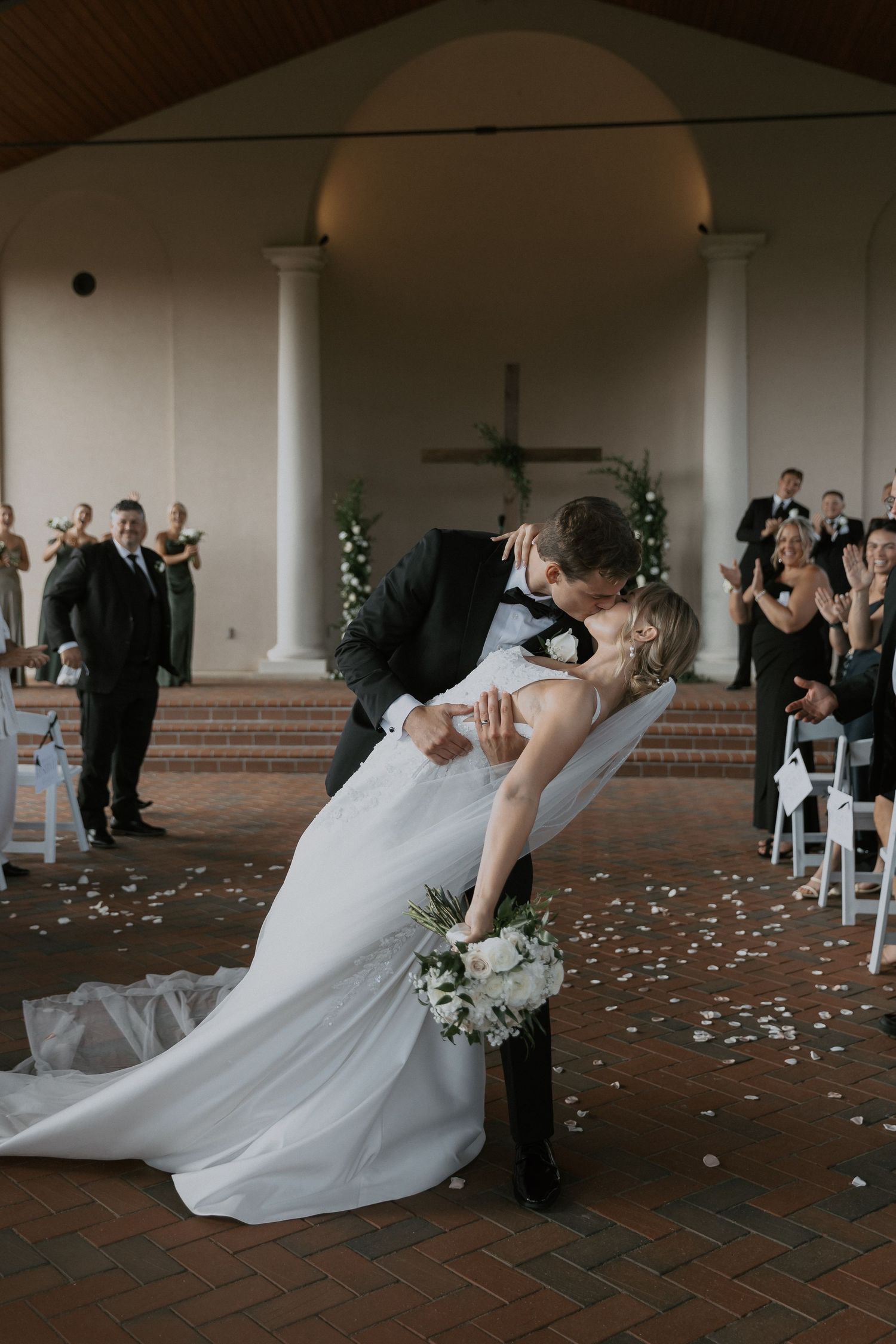 Wedding couple shares romantic dip kiss in chapel with guests applauding in Tuscany-inspired wedding venue.