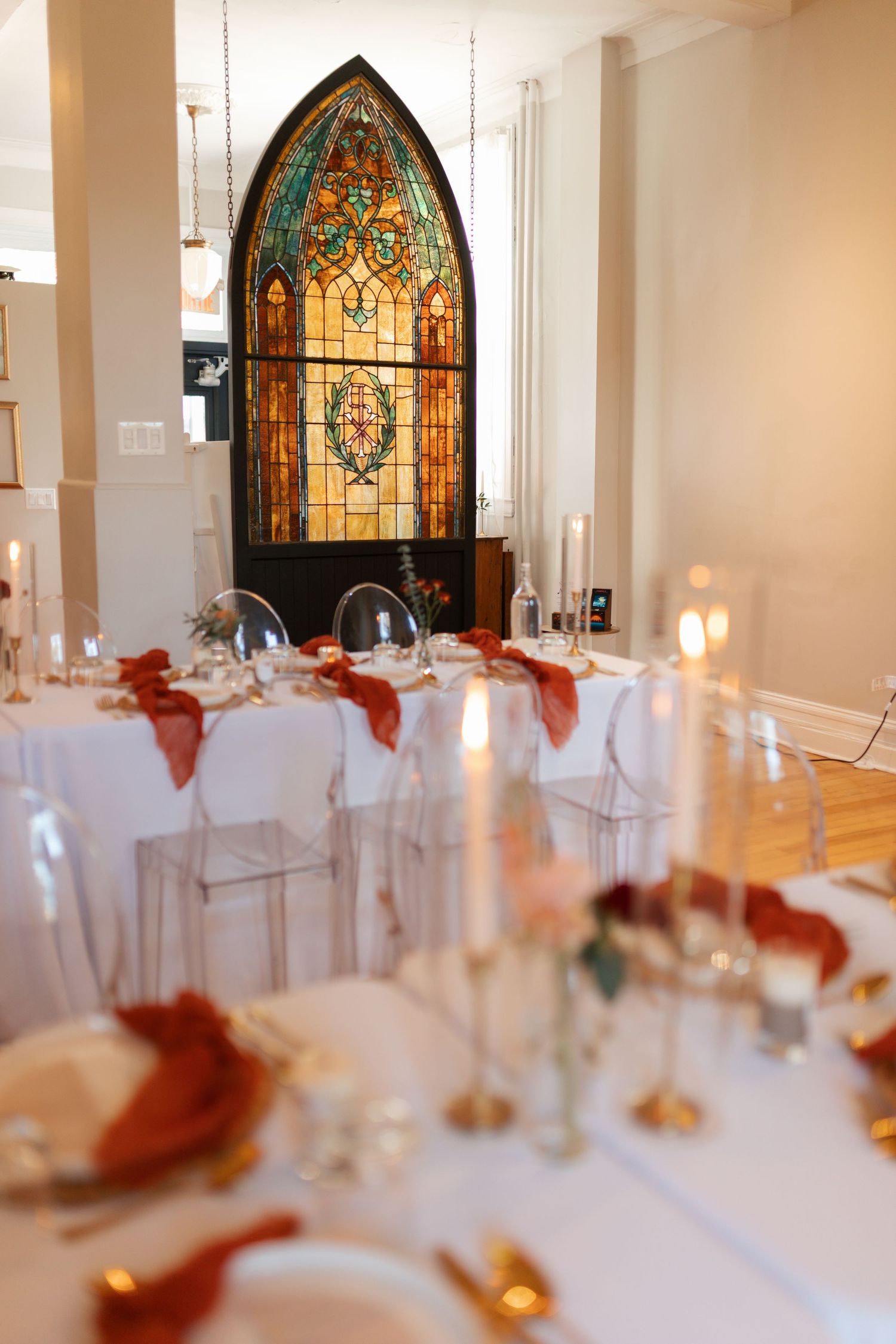 Elegant wedding reception hall with stained glass window, white tables, and rust-colored napkins.