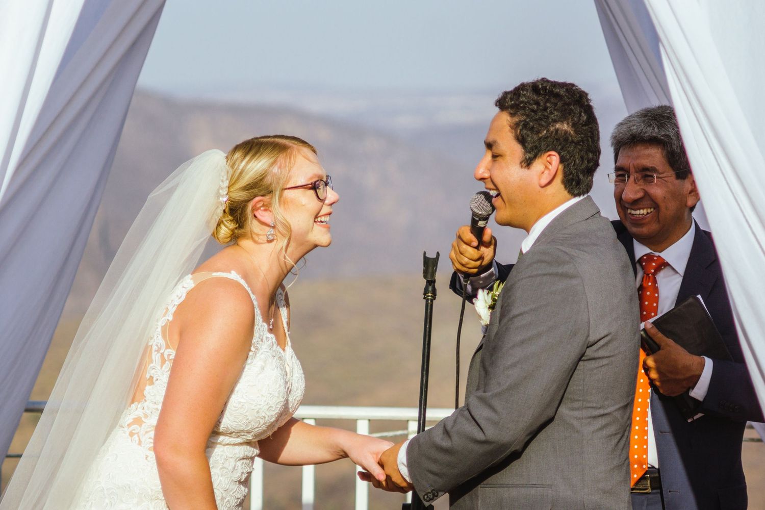 A couple shares a joyful moment at their outdoor wedding ceremony under a white canopy with mountains in the background.