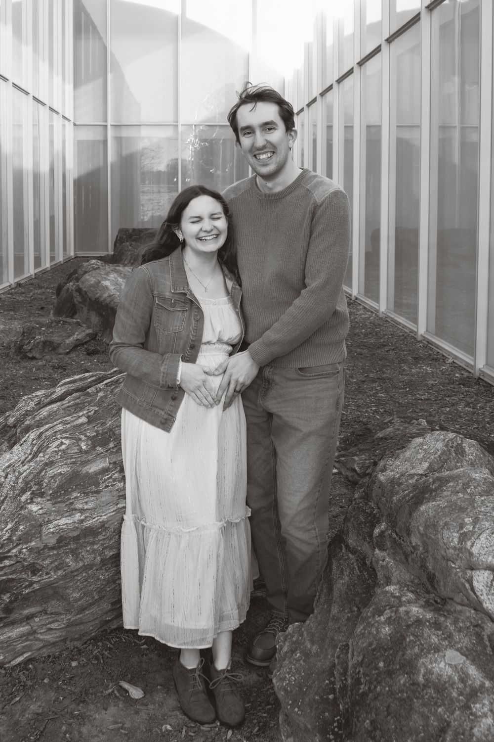 A couple stands together in front of a greenhouse on rocky ground, captured in black and white.