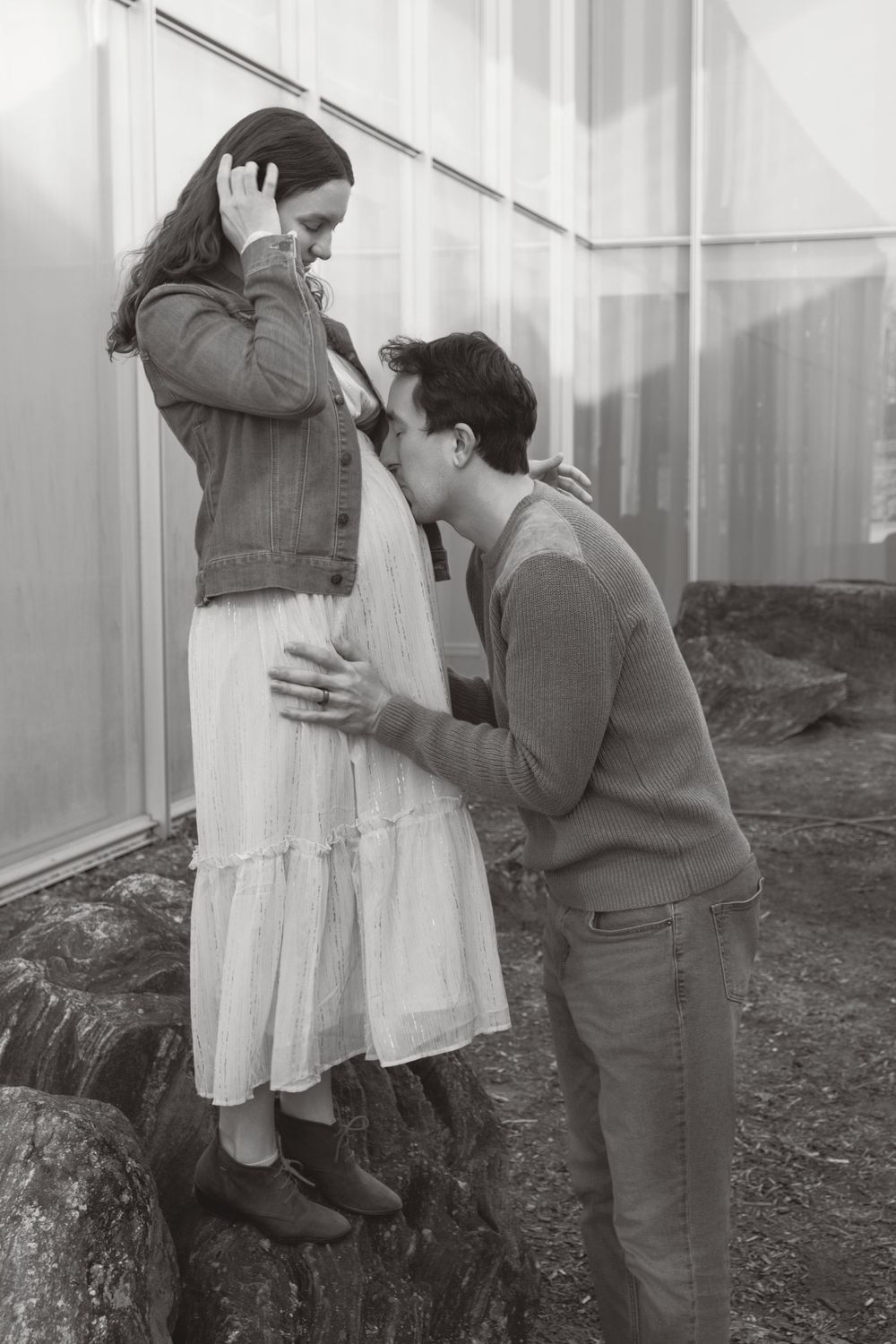 A romantic black and white portrait of a couple embracing outdoors near rocks, with her in a flowing dress and him in a sweater.