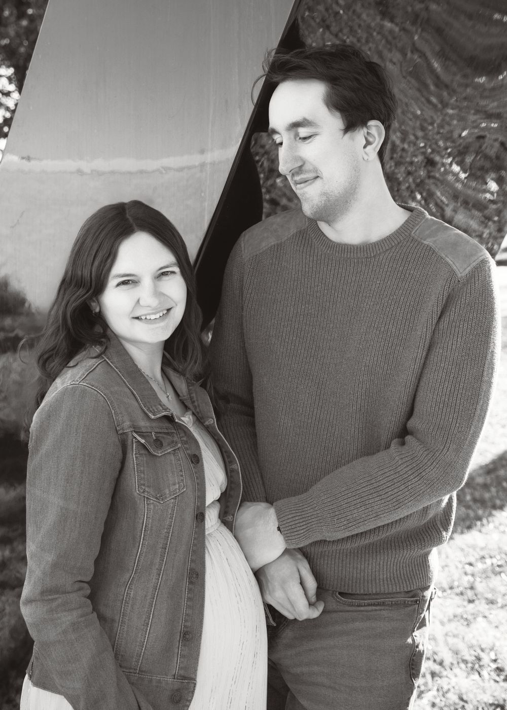 A casual black and white portrait of a couple smiling together outdoors, both wearing denim jackets and sweaters.
