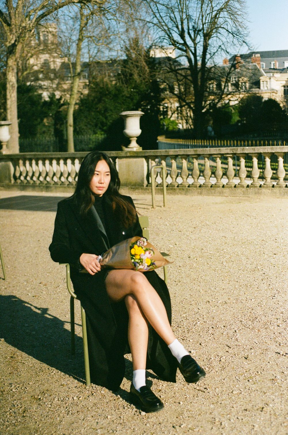 A person sits on a chair at a classic European garden terrace with ornate stone balustrades and trees in the background.