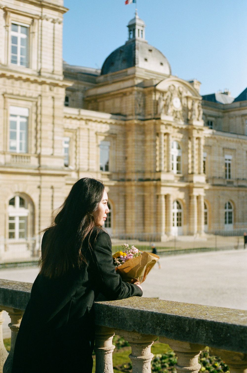 A person holding flowers stands on a terrace overlooking the Luxembourg Palace in Paris on a sunny day.