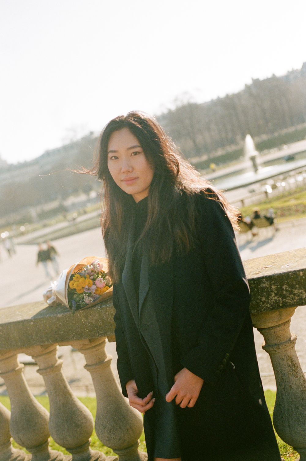 Person wearing a black winter coat poses on a stone balustrade during golden hour with trees in the background.