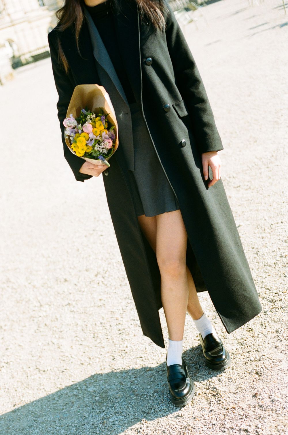 A person in a long black coat holding a bouquet of flowers walking outdoors wearing white socks and dark shoes.