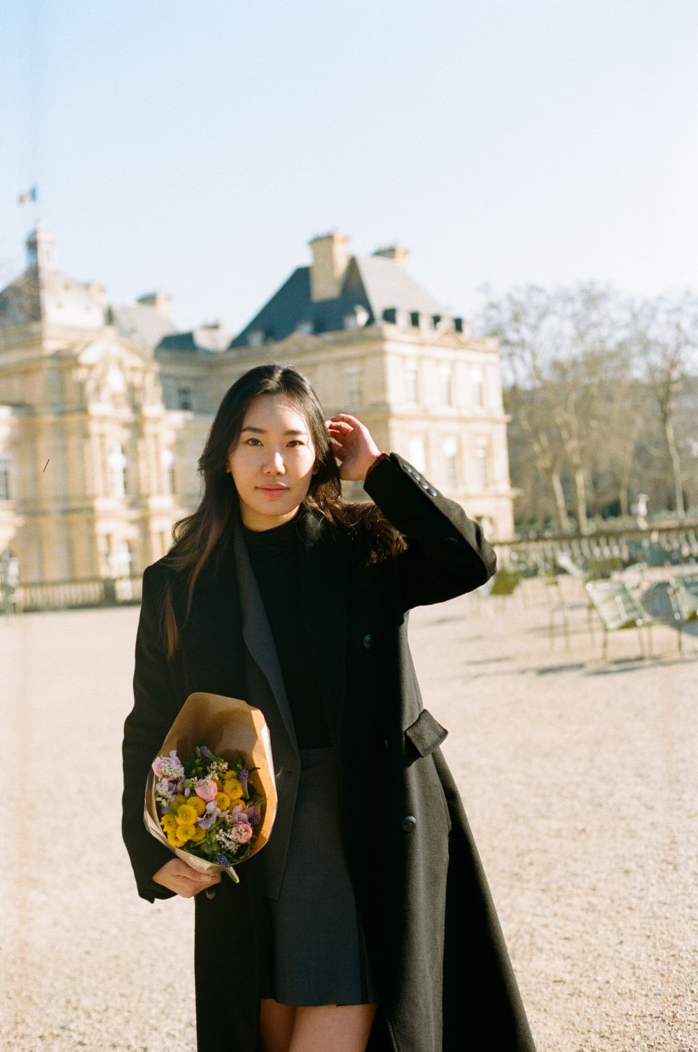 A person in a black coat holds a bouquet of flowers while posing outside the Luxembourg Palace.