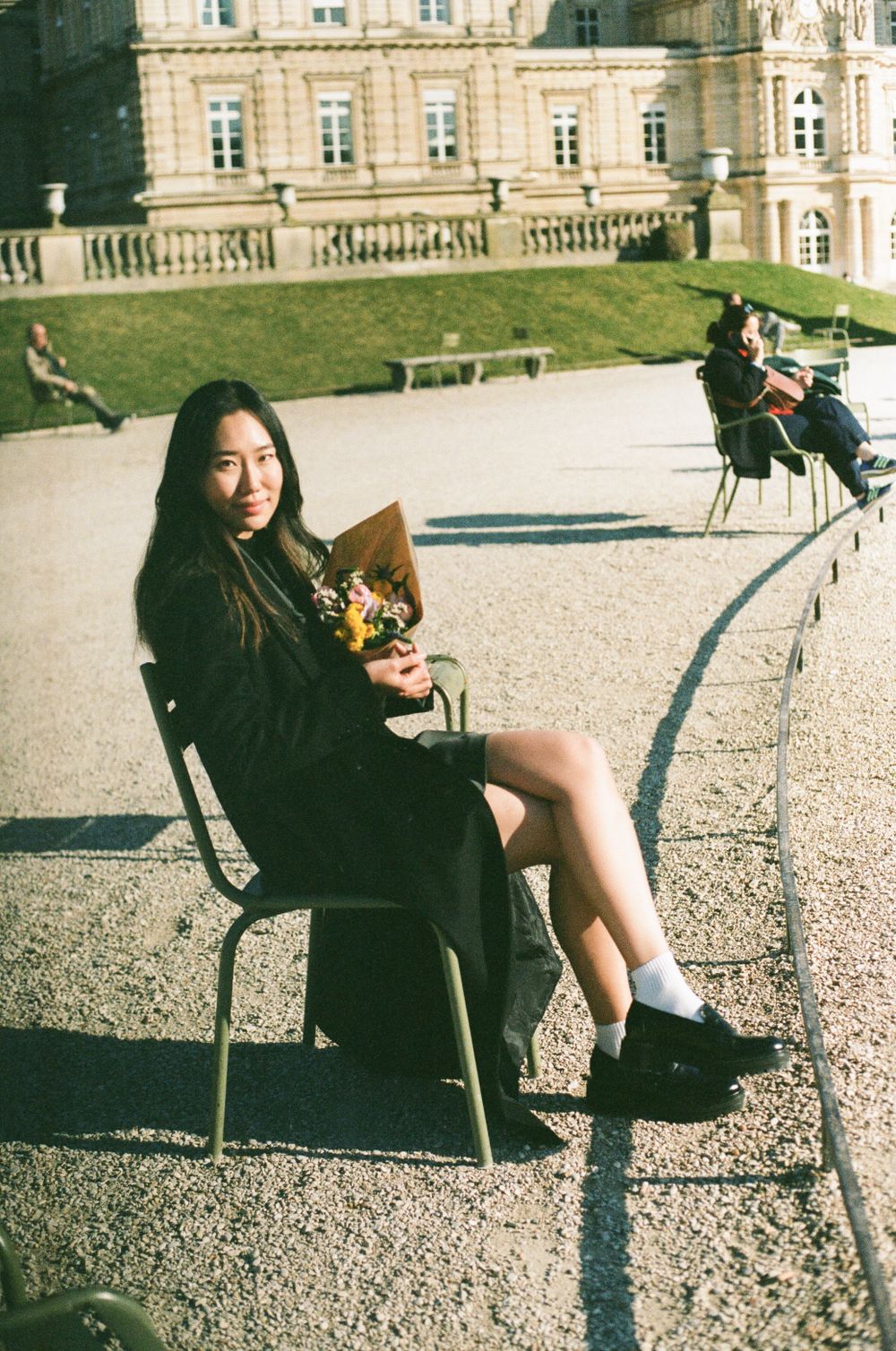 A person in a black dress sits on a chair holding flowers outside a grand European-style palace on a sunny day.