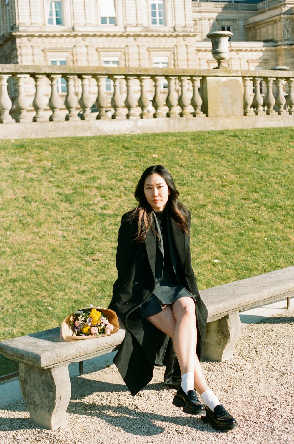 A person in a black coat sits on a stone bench near a classical building with ornate balusters and grassy lawn.