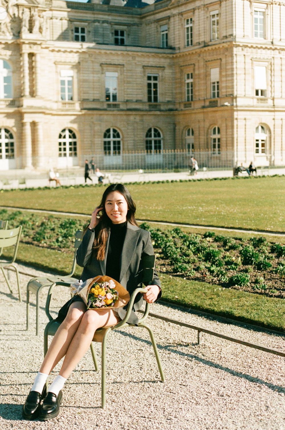 A stylish person relaxes in a chair on the grounds of an elegant French palace with manicured gardens.