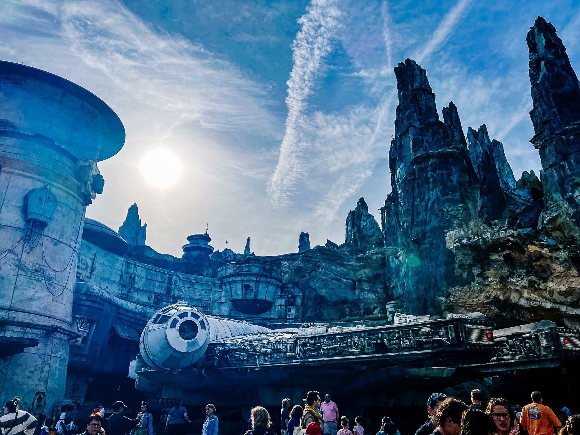 Crowds gather at Star Wars: Galaxy's Edge themed area with the Millennium Falcon spaceship and jagged rock formations under blue sky.