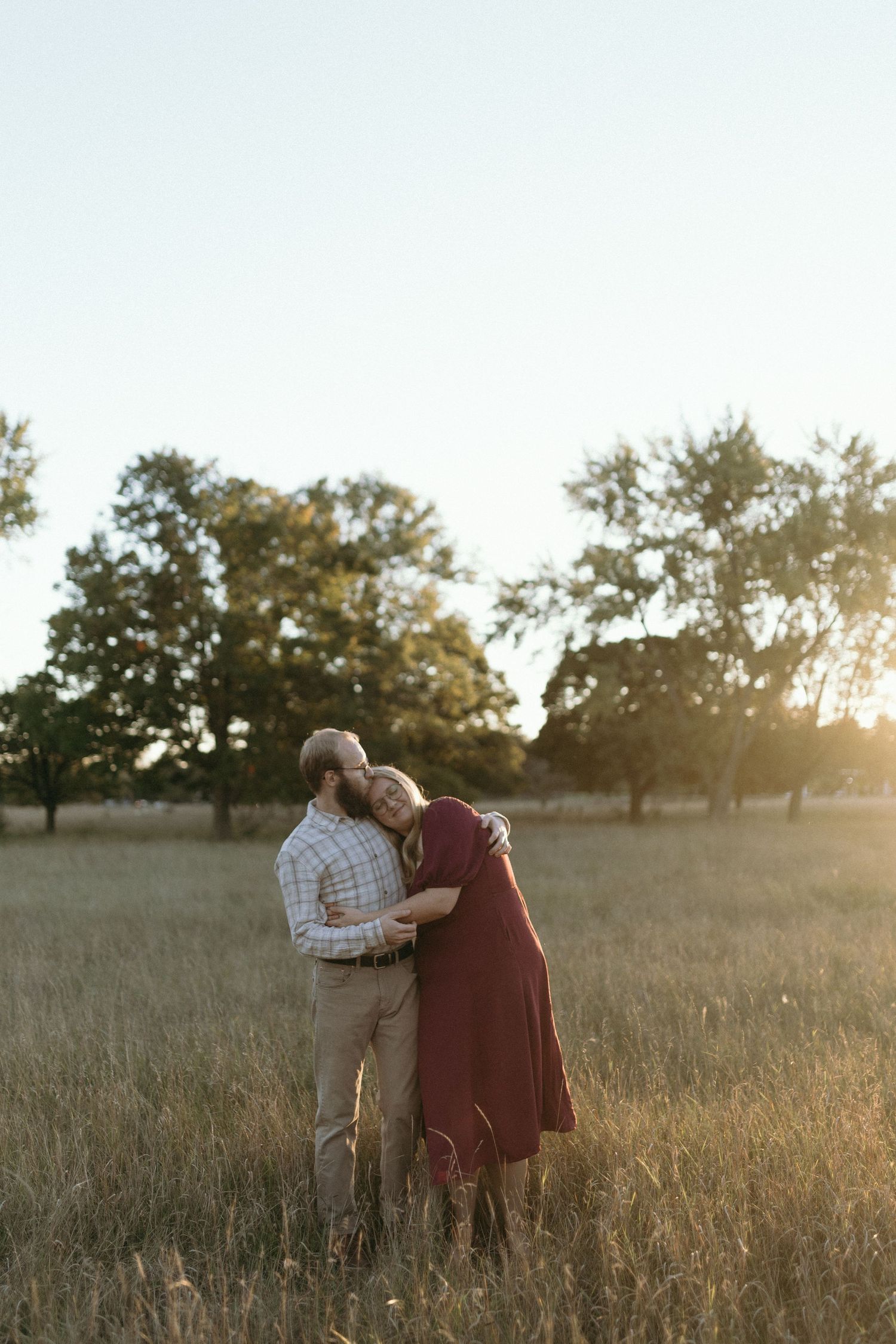 A couple embraces in a sun-drenched field at golden hour with tall grass and trees silhouetted in the background.