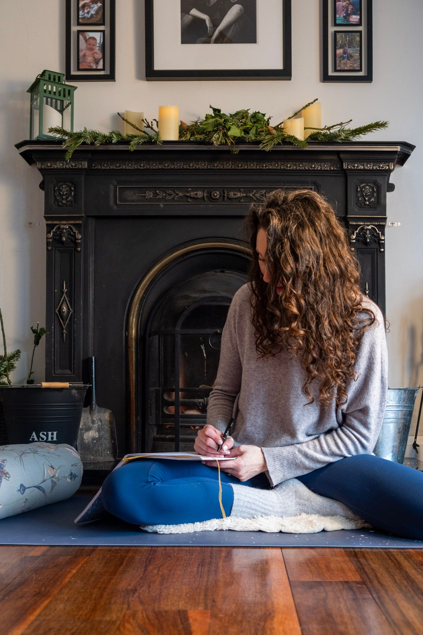 Reader sitting cross-legged by ornate black fireplace with holiday greenery on mantel.