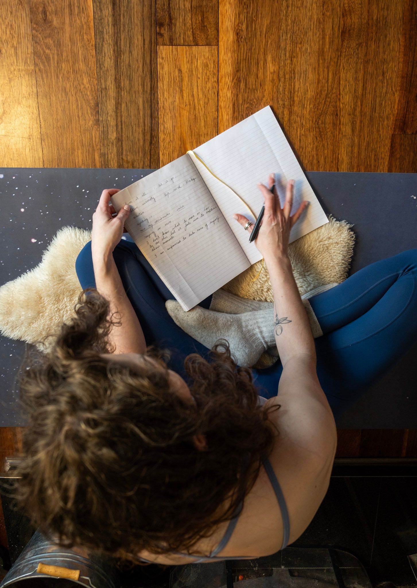 Person lies on wooden floor reading open book and writing and journaling on fuzzy white blanket.
