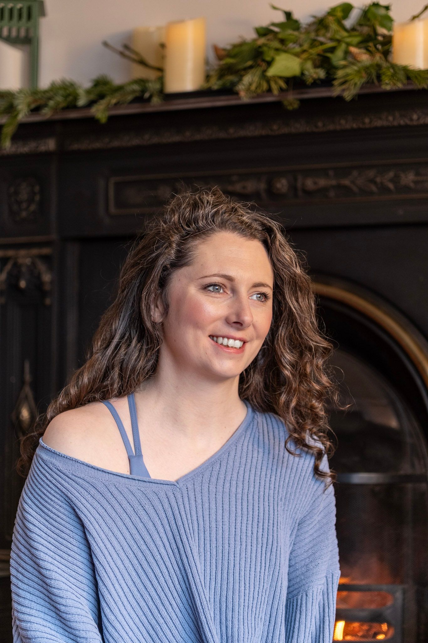 Woman in blue sweater sitting by decorated fireplace with greenery and candles.
