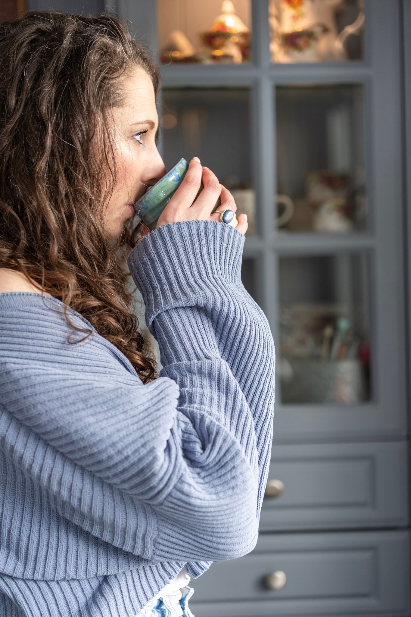 Person in grey sweater drinking from teal ceramic mug by cabinet.