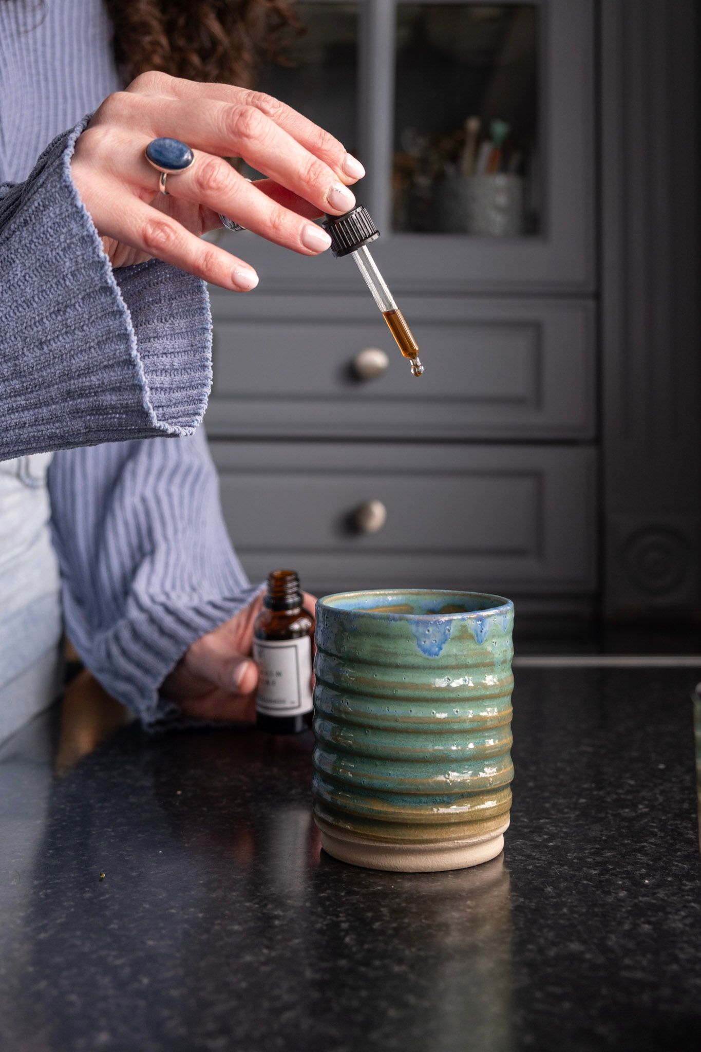 Hand holding dropper over turquoise ceramic mug with essential oil bottle on dark counter.
