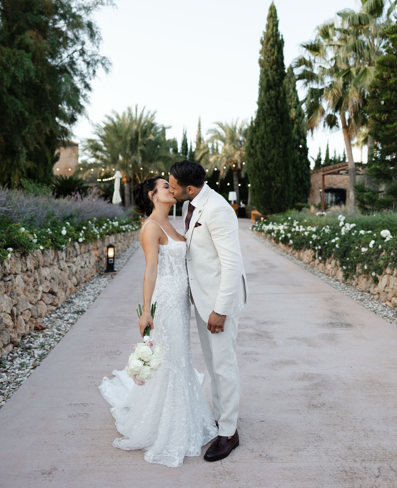 A couple shares a romantic moment on their wedding day along a garden path lined with cypress trees and flowers at their wedding in Spain.