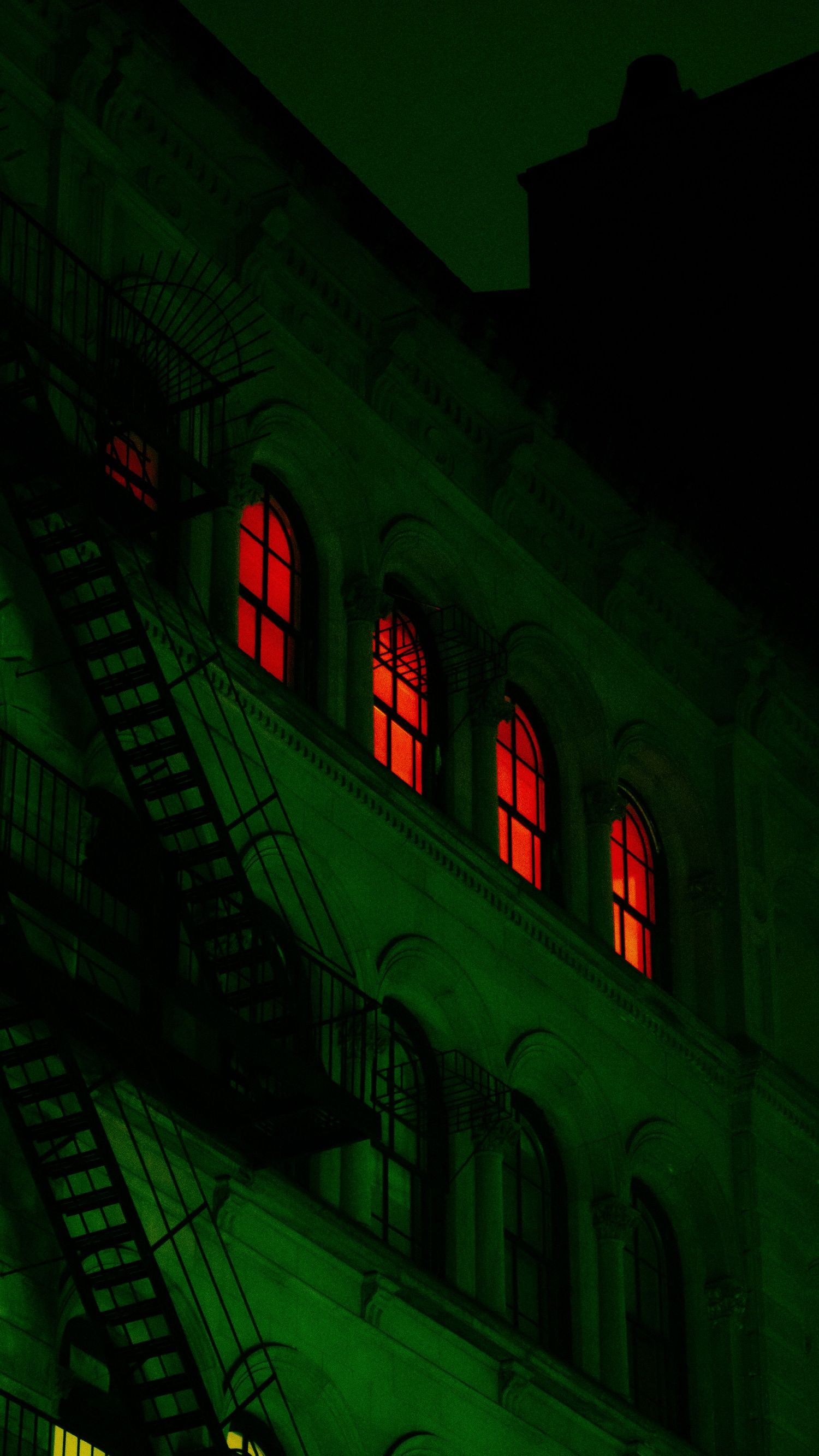 Urban building facade with red glowing windows against dark green lighting and fire escape silhouettes.