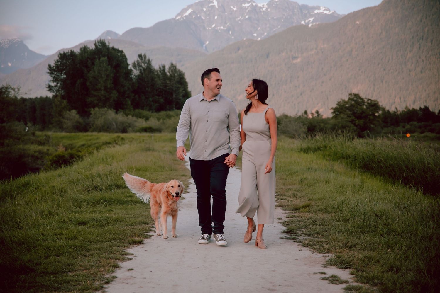 Couple walking with their golden retriever on a path with snow-capped mountains in the background.