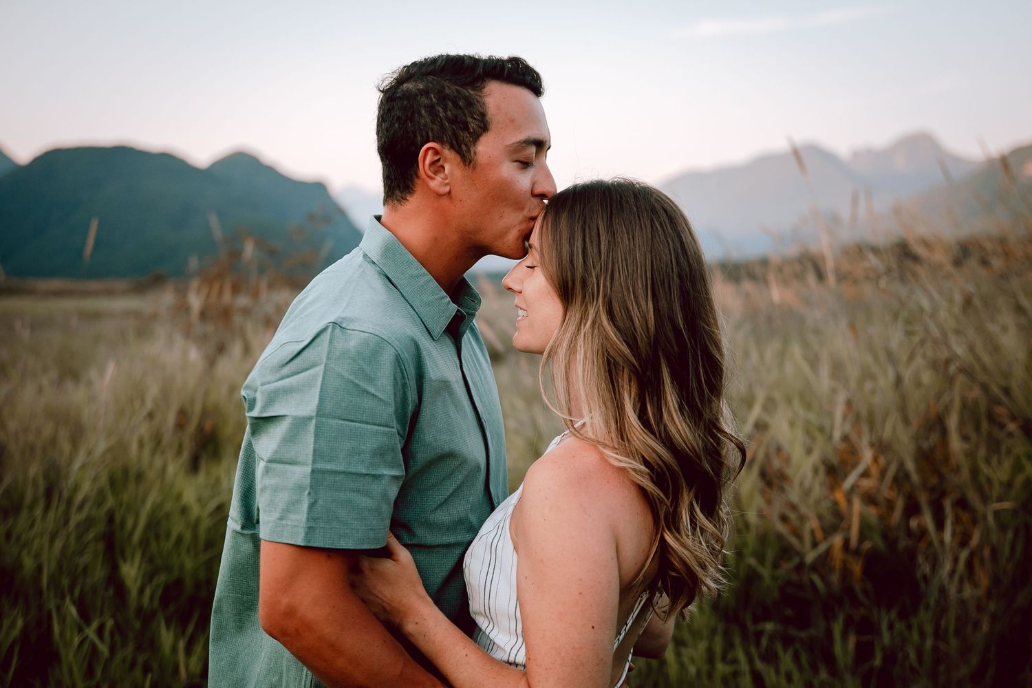 Couple embracing in field with mountain landscape at sunset.