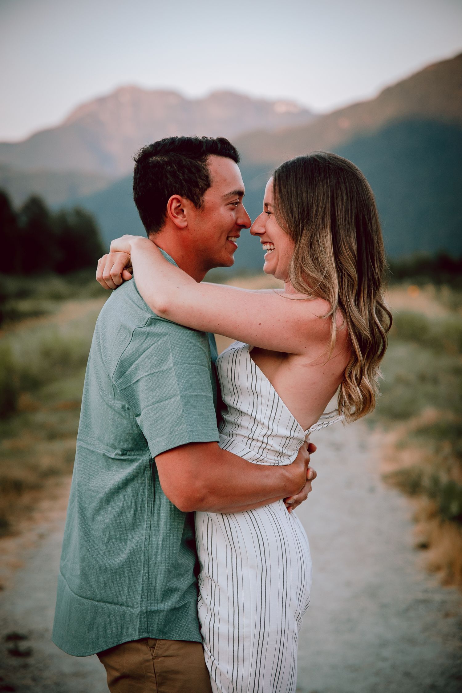A romantic couple embraces on a mountain trail at sunset while wearing casual summer outfits.