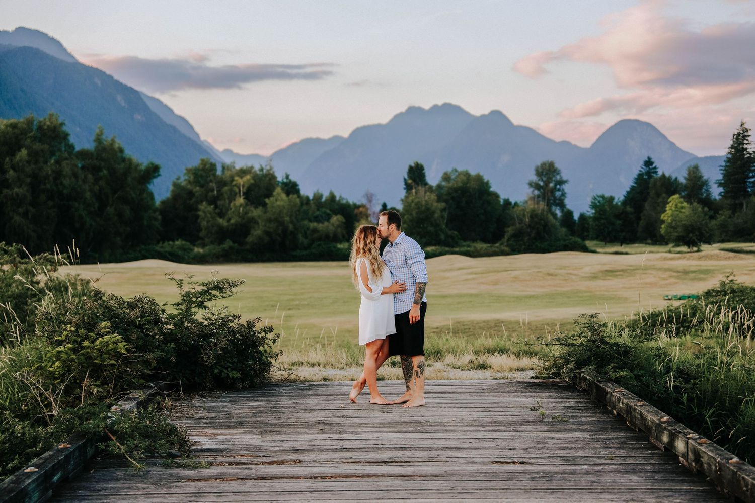 Couple sharing a romantic moment on a wooden boardwalk with mountain landscape in the background at sunset.