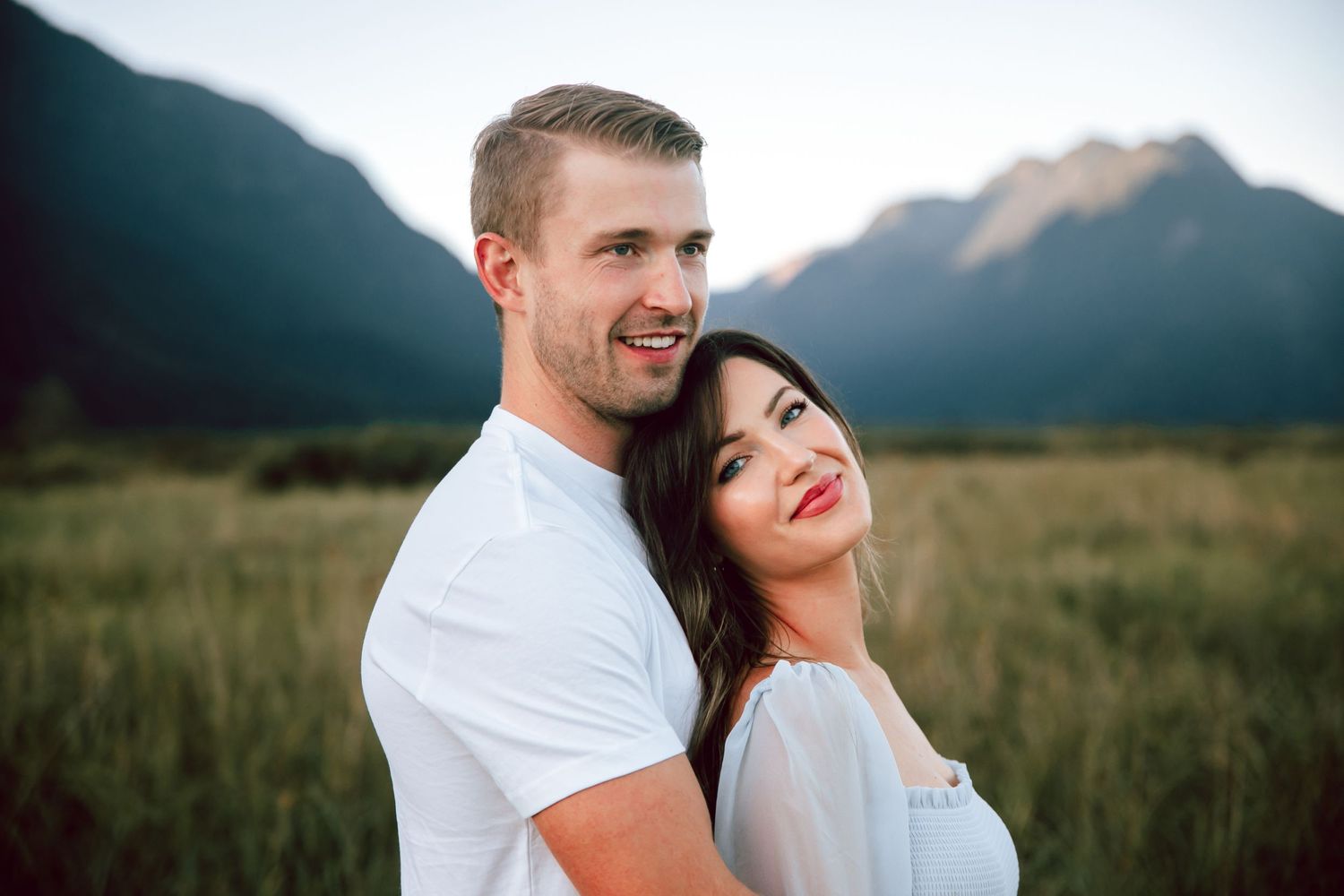 Happy couple smiles together with mountain peaks in background during sunset photoshoot.
