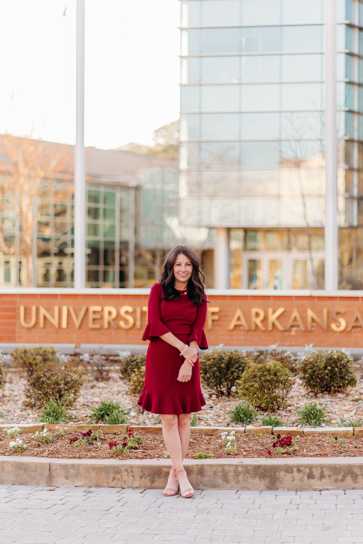 Woman in red dress poses elegantly at University of Arkansas campus entrance.
