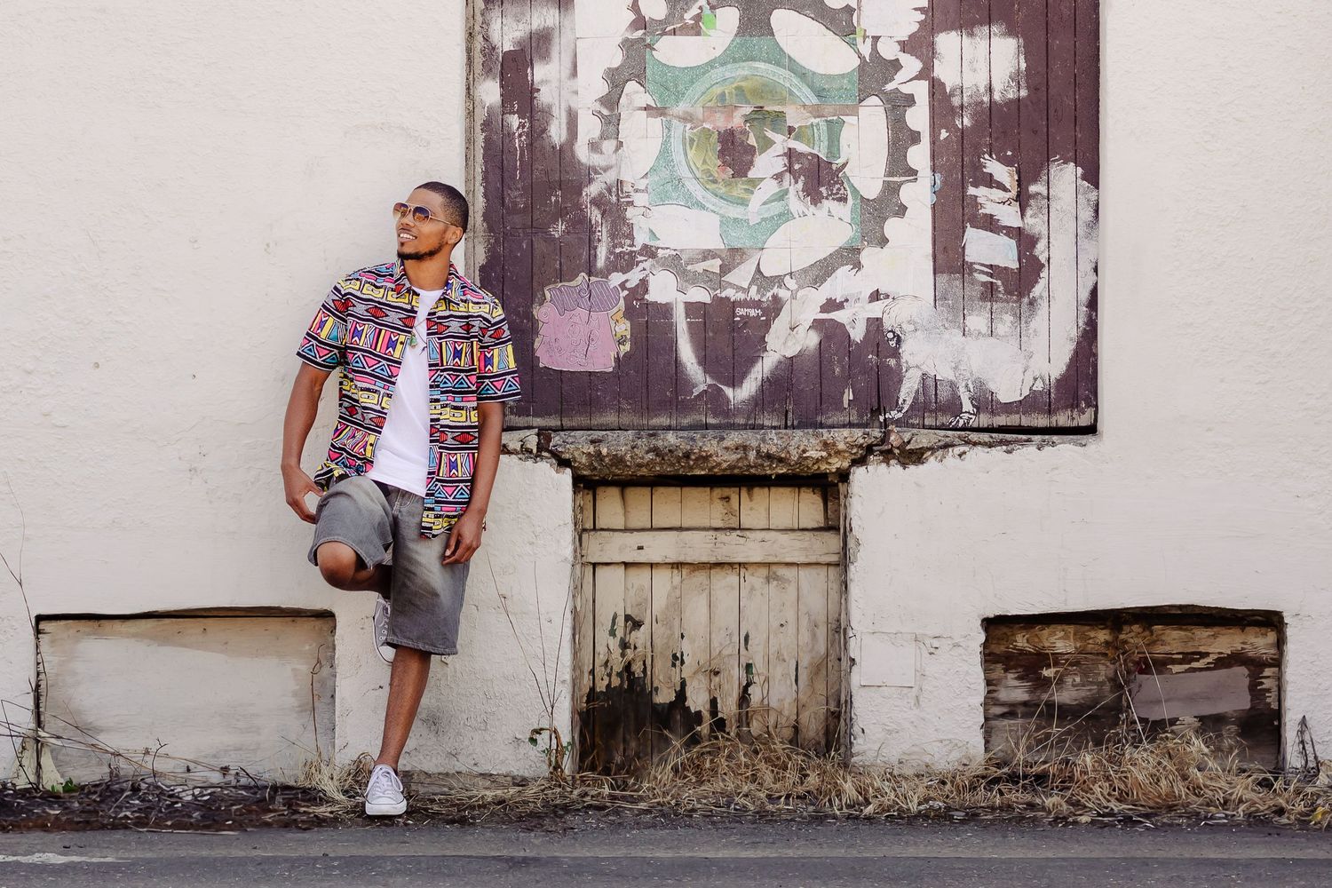 A person in a patterned shirt and shorts leans against a weathered white wall with graffiti art above.