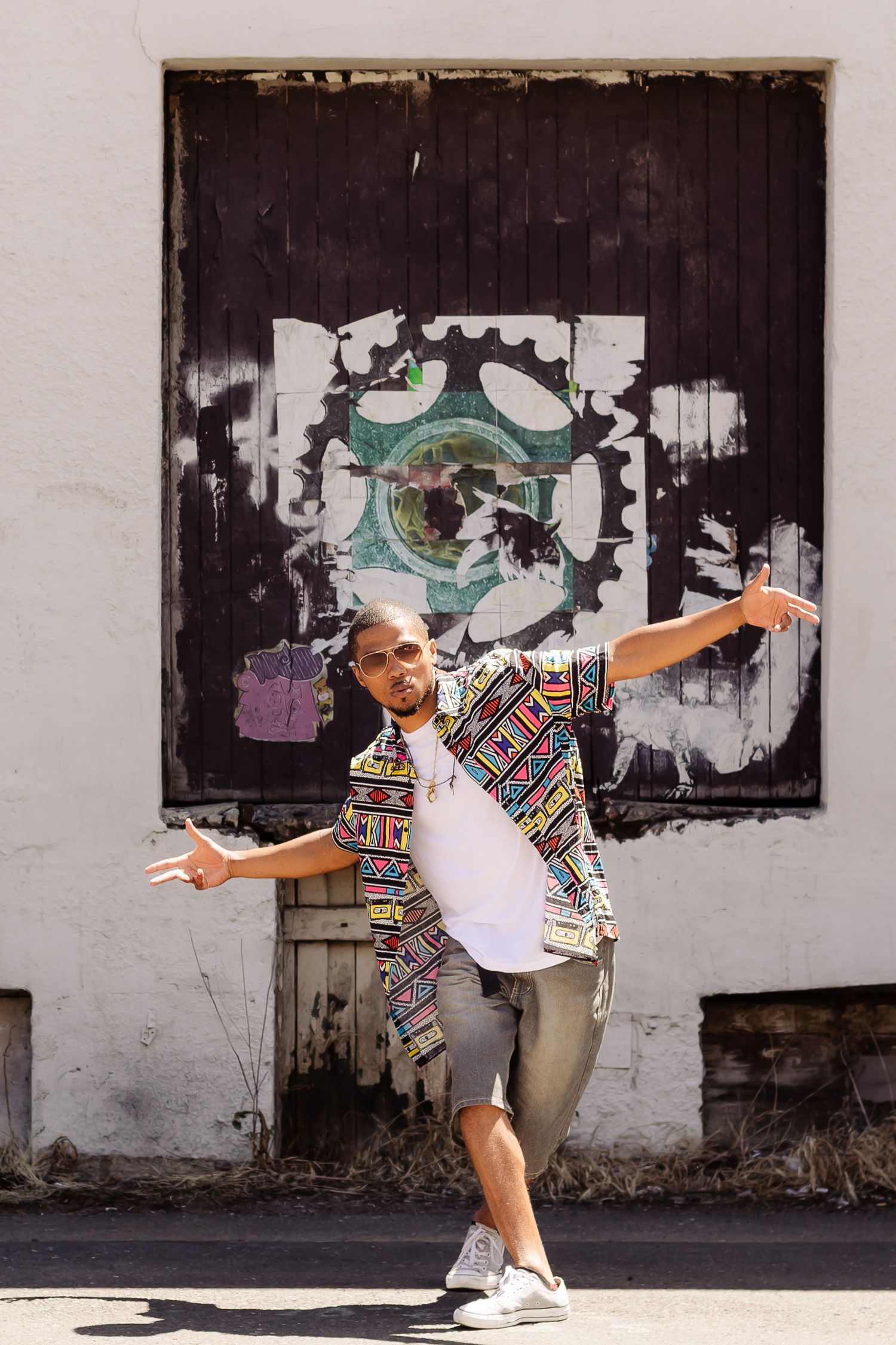 Person dancing joyfully on urban street wearing colorful patterned shirt and shorts against graffiti wall background.
