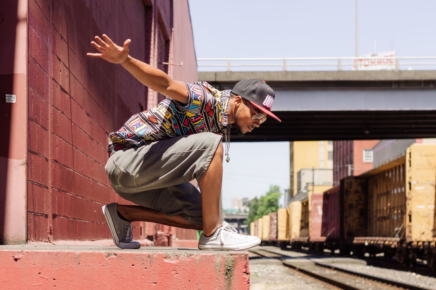 Urban dancer in colorful shirt performs dynamic crouching pose near train tracks with urban architecture backdrop.