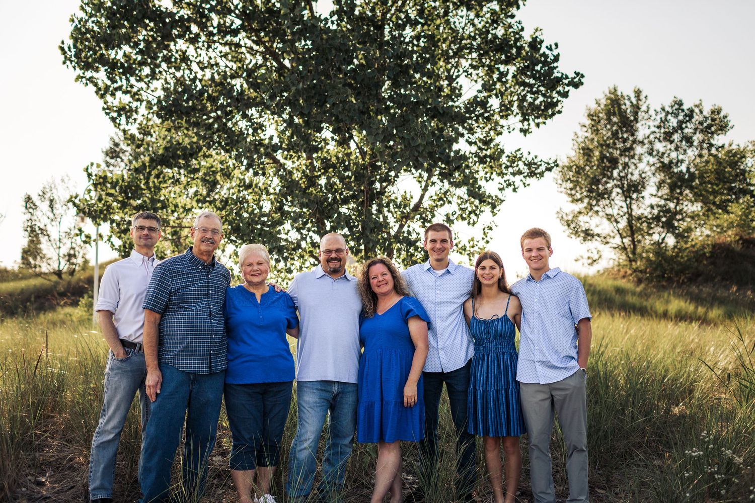 Family of eight smiling together outdoors in a grassy field with a large tree backdrop during golden hour.