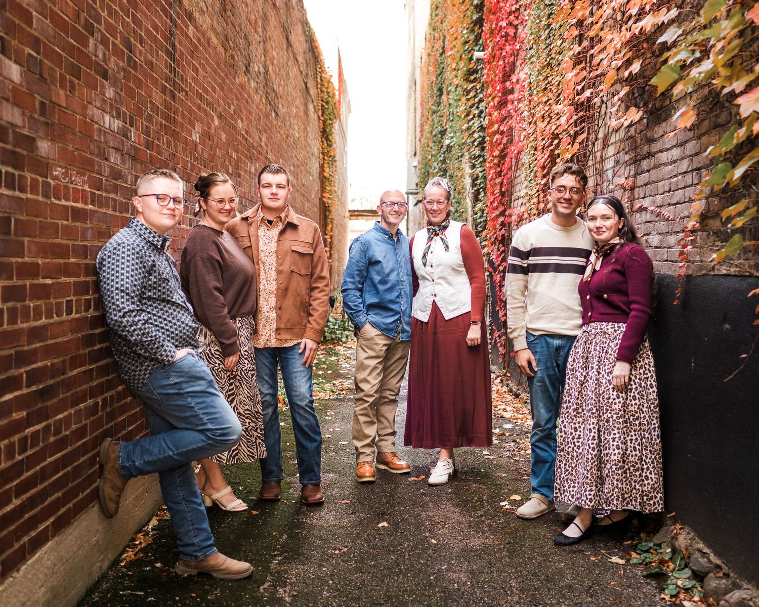 Group of seven people smiling in a brick alley with colorful autumn ivy, dressed in casual fall outfits.