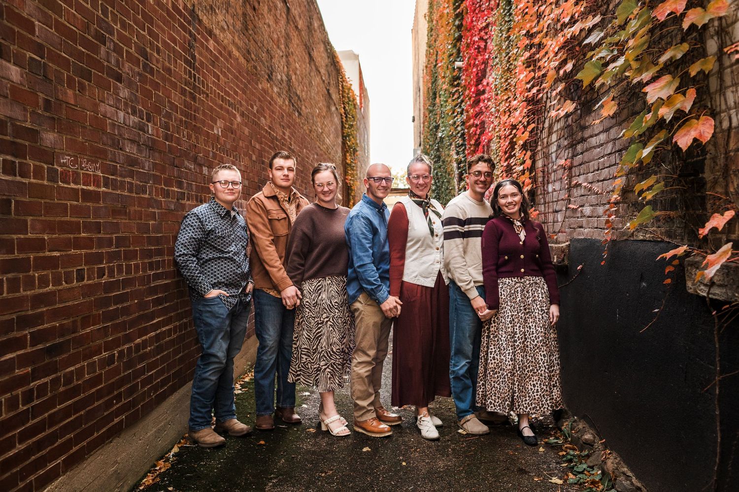 Family of seven posing in a brick alley with colorful autumn ivy, smiling together in fall attire.